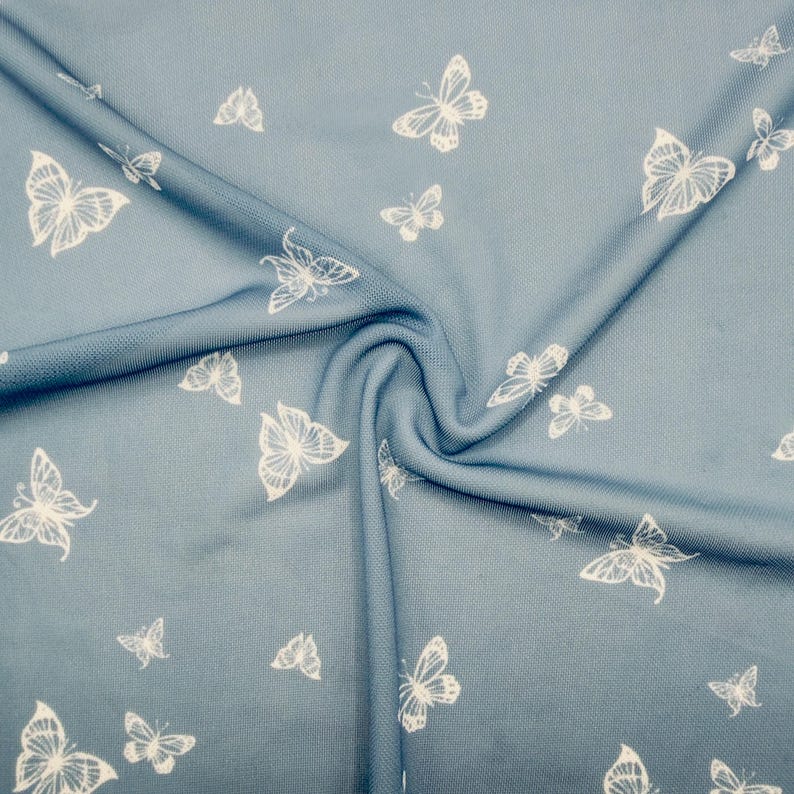 May include: A close-up of a blue fabric with a white butterfly pattern. The butterflies vary in size and are scattered across the material. The fabric is gathered in a swirl, showing the texture and folds of the material.