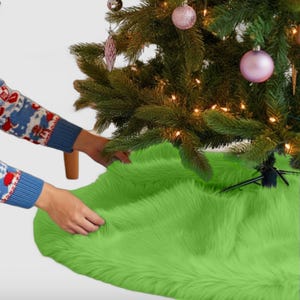 May include: A bright green faux fur Christmas tree skirt. The skirt is being placed under a Christmas tree by a person's hand.