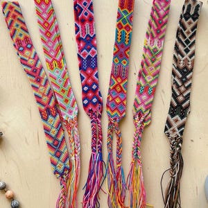 Può includere: Sei braccialetti dell'amicizia tessuti colorati con intricati motivi geometrici. Ogni braccialetto presenta una diversa combinazione di colori, tra cui vibranti tonalità arcobaleno, rosa, blu e marrone. Le estremità sono rifinite con nappe intrecciate di fili coordinati.