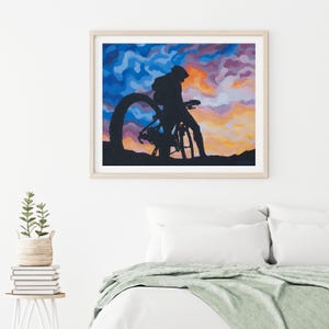 K&ouml;nnte beinhalten: Gerahmter Kunstdruck mit einer Silhouette einer Person mit Fahrrad vor einem farbenfrohen Sonnenuntergang. Das Kunstwerk zeigt einen Radfahrer und ein Fahrrad in Schwarz, vor einem lebendigen Himmel aus Blau, Orange und Lila. Der Druck ist in einem hellen Holzrahmen gefasst.