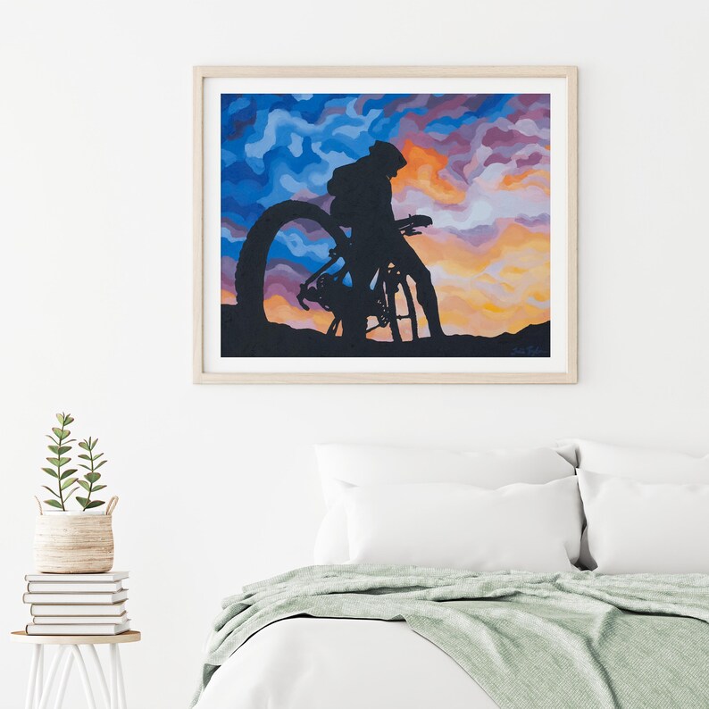 K&ouml;nnte beinhalten: Gerahmter Kunstdruck mit einer Silhouette einer Person mit Fahrrad vor einem farbenfrohen Sonnenuntergang. Das Kunstwerk zeigt einen Radfahrer und ein Fahrrad in Schwarz, vor einem lebendigen Himmel aus Blau, Orange und Lila. Der Druck ist in einem hellen Holzrahmen gefasst.