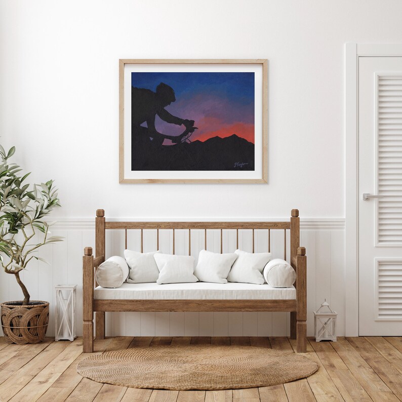 May include: Framed artwork showing a cyclist silhouetted against a sunset. The artwork depicts a cyclist riding a bicycle on a mountain, with a gradient sky of blue, purple, and orange hues. The artwork is in a wooden frame.