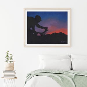 May include: Framed art print showing a cyclist silhouetted against a sunset. The artwork depicts a cyclist on a mountain road with a blue, purple, and red sky. The print is in a light-coloured wooden frame, suitable for a bedroom or living room.