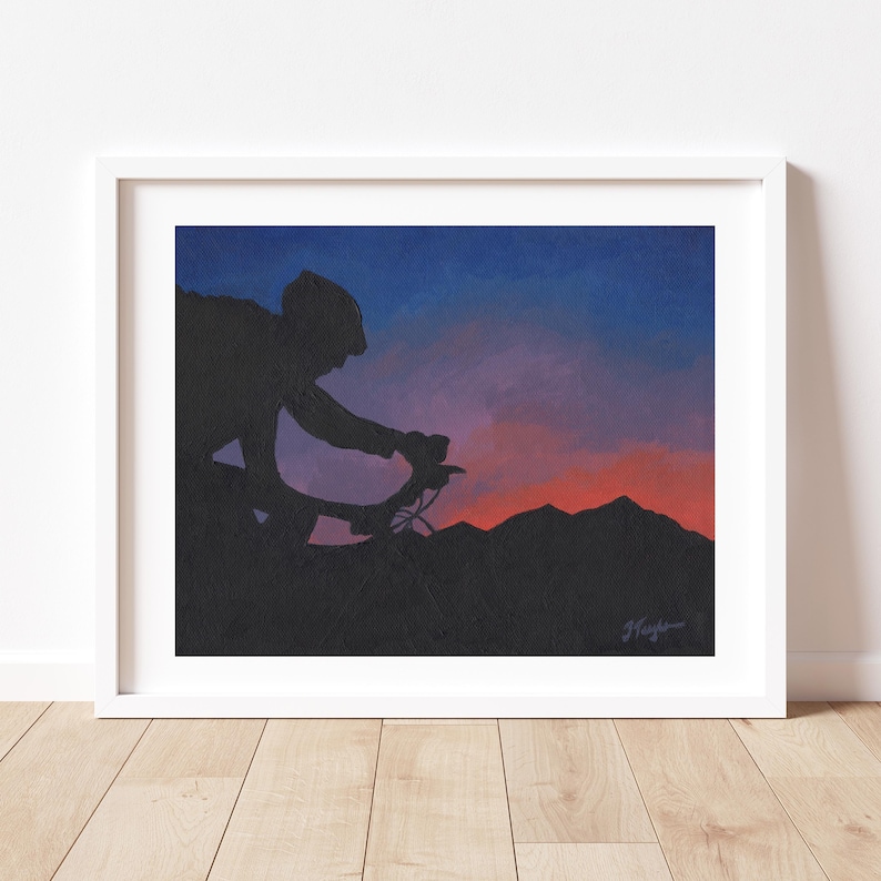 May include: Framed artwork depicting a cyclist silhouetted against a sunset. The sky transitions from blue to orange and red, with dark mountain silhouettes. The artwork is in a white frame. The image evokes a sense of movement and adventure.