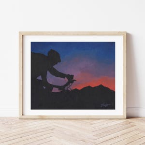 May include: Framed art print depicting a cyclist's silhouette against a sunset. The artwork shows a cyclist on a bicycle with mountains in the background. The sky transitions from dark blue to red and orange.