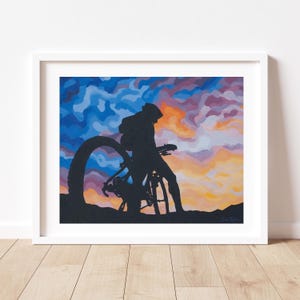 K&ouml;nnte beinhalten: Gerahmter Kunstdruck mit einer Silhouette eines Radfahrers und Fahrrads vor einem farbenfrohen Sonnenuntergangshimmel. Das Kunstwerk befindet sich in einem wei&szlig;en Rahmen und wird auf einem Holzboden pr&auml;sentiert. Die Farbpalette umfasst Blau-, Orange- und Lilat&ouml;ne.