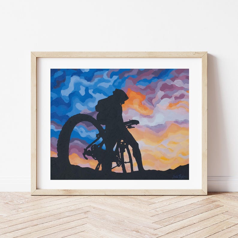 K&ouml;nnte beinhalten: Gerahmter Kunstdruck mit einer Silhouette eines Radfahrers und Fahrrads vor einem farbenfrohen Sonnenuntergangshimmel. Das Kunstwerk befindet sich in einem Holzrahmen und wird an einer wei&szlig;en Wand und einem Holzboden gezeigt.