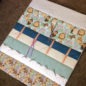 May include: A multi-colored baby play mat with a jungle animal theme. The mat has different textures and patterns, including a white fabric with grey polka dots, a blue fabric with white stripes, and a beige fabric with orange polka dots. The mat also has several loops and ties for attaching toys.