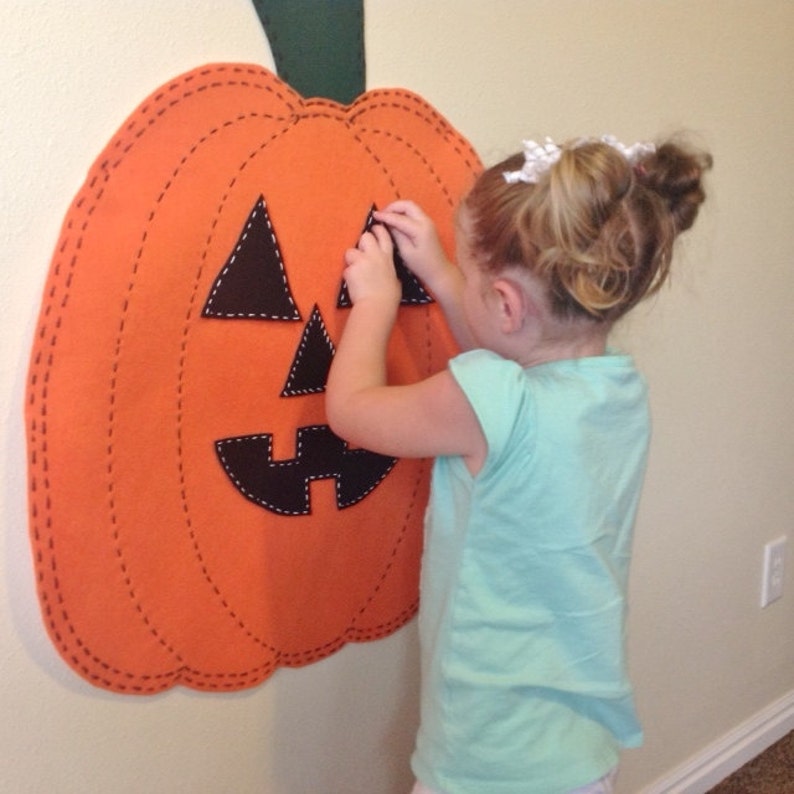 May include: A child places felt pieces on a large orange felt pumpkin with a stitched outline. The pumpkin has a black felt face with a triangle nose, two eyes, and a wide smile.