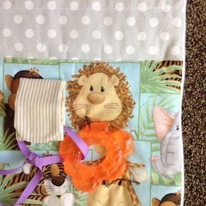 May include: A baby activity play mat with a variety of textures and colors. The mat features a lion, an elephant, a tiger, and a giraffe. There is also a teething ring and a sock attached to the mat.