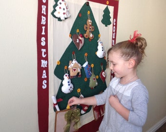 May include: A felt Christmas countdown advent calendar with a green tree and various felt ornaments. A young girl is opening a pocket on the calendar.