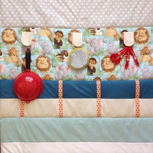May include: A multi-colored fabric play mat with a jungle animal print. The mat has five pockets with various items hanging from them, including a red colander, a yellow and gray textured disc, and red measuring spoons.