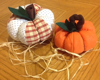 May include: Two handmade fabric pumpkins, one with a red and white plaid pattern and white polka dots, and the other a solid orange. Both pumpkins have green felt leaves and brown felt stems.