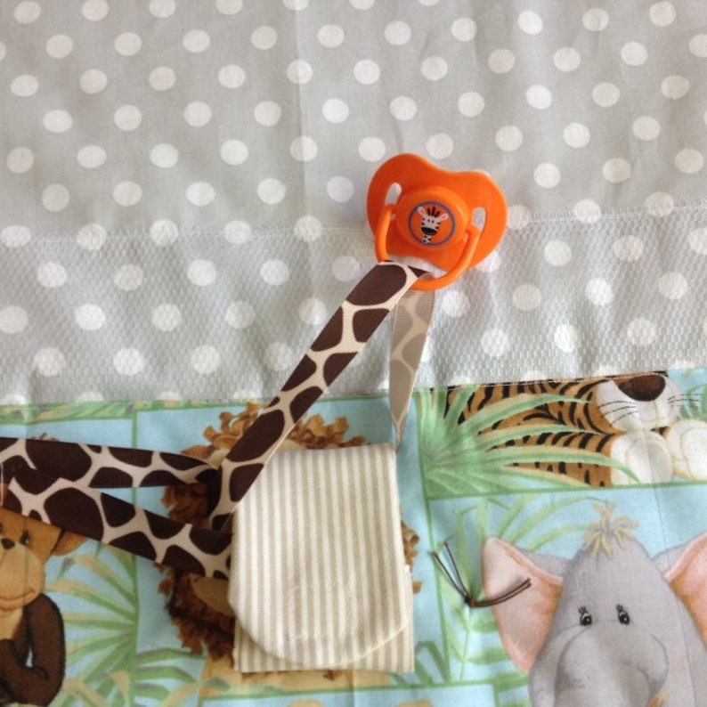 May include: A brown and white striped fabric pouch with a brown and orange giraffe print pacifier clip attached. The clip has a pacifier with a giraffe face on it. The pouch and clip are on a multi-colored fabric background with a tiger, elephant, and giraffe print.