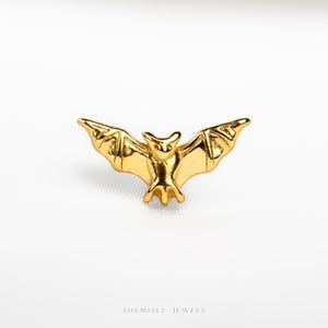 May include: A gold-toned metal stud earring shaped like a bat with outstretched wings. The bat features detailed wing structures and a pointed ear design. Below the bat, the text SHEMISLI JEWELS is printed in a small, clear, sans-serif font.