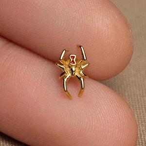 May include: A small, gold-colored spider charm with a red hourglass marking on its back. The spider has eight legs and is highly detailed. The charm is held between two fingers.