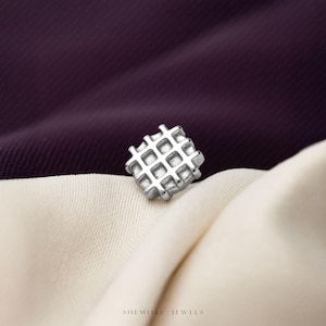 May include: A small, square silver stud earring featuring a raised grid or waffle pattern on its surface. The metal has a matte, textured finish, and the piece is positioned centrally. The text SHEMISEI JEWELS is printed at the bottom of the image.