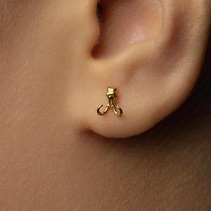 May include: A small, gold-colored earring with a unique design, featuring a central post and two curved, open loops. The earring is set against the backdrop of a person's ear and skin.