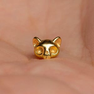 May include: A small, polished gold-toned metal stud earring shaped like a cat head. The design features two pointed ears, two smooth circular indentations for eyes, and a small triangular nose detail, all rendered in a reflective metallic finish.