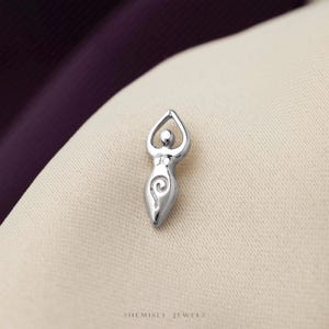 May include: A polished silver-toned pendant shaped like a stylized human figure with arms raised above the head in a triangular form and a spiral design engraved on the lower torso. The piece features smooth, reflective surfaces and clean lines.