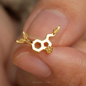 May include: A gold-colored stud earring shaped like a chemical molecular structure. The design features a hexagonal ring, a pentagonal ring, and a small flower detail at one end, with a leaf-like shape extending from the opposite side.