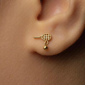 May include: A gold-colored tennis racket and ball earring. The earring is a small, detailed charm of a tennis racket with a ball hanging below. The earring is attached to an ear.