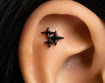 May include: A black metal ear piercing jewellery piece featuring a cluster of three four-pointed star shapes. The jewellery is positioned in the upper cartilage area of the ear, showcasing a smooth, dark finish and a distinct, sharp-edged star design.