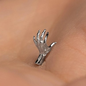 May include: A silver-colored hand-shaped jewelry piece. The hand is sculpted with detailed fingers and a visible wrist. The jewelry is likely a brooch, pendant, or earring, and is set against a neutral background.