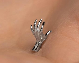 Può includere: Un gioiello a forma di mano color argento. La mano è scolpita con dita dettagliate e un polso visibile. Il gioiello è probabilmente una spilla, un ciondolo o un orecchino, su sfondo neutro.