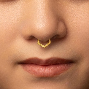 May include: A gold-toned septum ring featuring a V-shaped design with a textured, ribbed surface. The jewelry is positioned in the center of the nose, showcasing a metallic finish and a geometric chevron-inspired shape that follows the nasal contour.