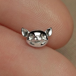 May include: A small, silver-toned metal stud earring shaped like a pig head. The design features pointed ears, two circular eyes, a snout with two nostrils, and a curved mouth line, all rendered in a polished, reflective metallic finish.
