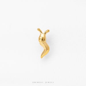 May include: A small, gold-toned metal stud earring shaped like a stylized snail with two antennae and a curved body. The piece features a smooth, polished finish and is positioned above the text SHEMISLI JEWELS printed in a simple font.