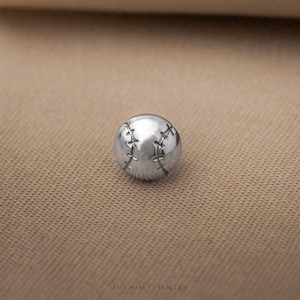 May include: A small, spherical silver-toned charm shaped like a baseball, featuring detailed engraved stitching lines across its surface. The object has a polished metallic finish and is positioned centrally, with the text SHEMISEI JEWELS visible below.
