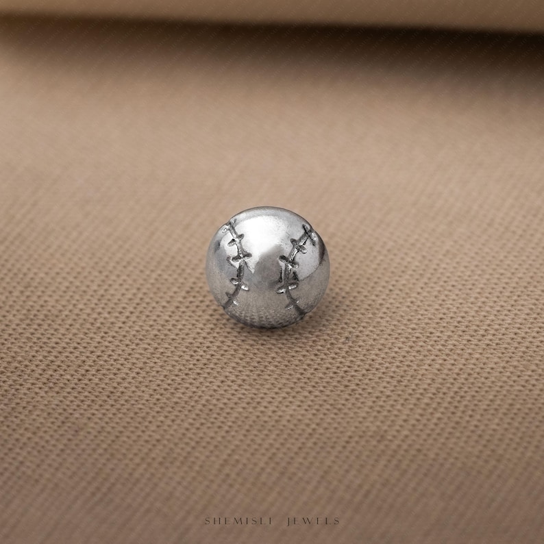 May include: A small, spherical silver-toned charm shaped like a baseball, featuring detailed engraved stitching lines across its surface. The object has a polished metallic finish and is positioned centrally, with the text SHEMISEI JEWELS visible below.