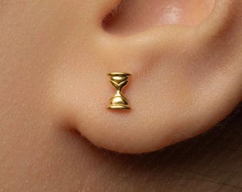 May include: A small gold stud earring shaped like an hourglass. The earring features a polished, reflective metallic finish with a narrow centre and wider top and bottom sections, securely positioned on the lower part of an earlobe.