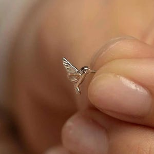 May include: A small silver-toned hummingbird stud earring featuring detailed wing and body engravings. The bird is depicted in a flying position with its wings angled upward and a pointed beak extending forward, showcasing a polished metallic finish.