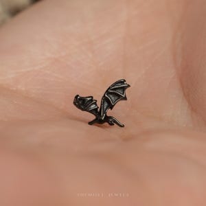 May include: A small, dark metallic stud earring shaped like a dragon with outstretched wings. The piece features detailed, textured wing membranes and a pointed tail. The text 'SHEMISLI JEWELS' is printed in small white letters at the bottom center.