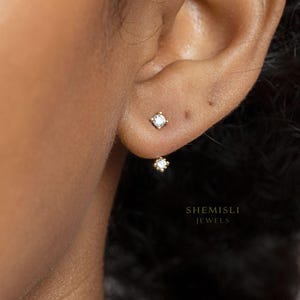 May include: A close-up of a person's ear adorned with two gold earrings. The top earring is a small square shape with a clear stone. The second earring is a small flower shape with a clear stone. The text "SHEMISLI JEWELS" is visible.