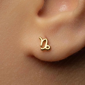 May include: A small gold stud earring shaped like the Capricorn zodiac symbol. The earring features a polished, smooth finish and is designed to sit flush against the earlobe, showcasing the distinct curved lines of the astrological sign.