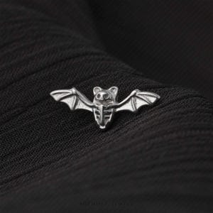 May include: A silver-toned metal pin shaped like a bat with outstretched wings. The bat features a detailed skeletal chest design and pointed ears. The surface of the metal has a polished finish with recessed areas that highlight the bat's anatomy.