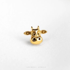 May include: A small, polished gold-tone stud earring shaped like a cow head, featuring two ears, two eyes, and a snout with two nostrils. The text SHEMISLI JEWELS is printed in small, dark letters at the bottom of the frame.