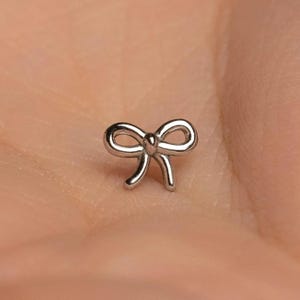 May include: A small, silver-colored bow-shaped stud earring. The bow has a classic design with looped ends and a central knot. The earring is shown against a blurred background.