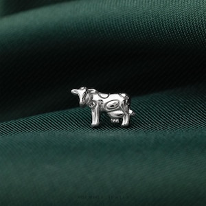 May include: A small, silver-toned metal stud earring shaped like a cow. The cow figure features a smooth, reflective surface with embossed circular spots across its body and head. The text "SILIMISLI JEWELS" is printed in small letters at the bottom.