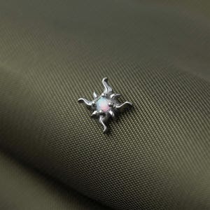May include: A silver-toned metal stud earring featuring a central round iridescent opal stone. The setting is shaped like a four-pointed star with curved, flame-like rays extending outward from the center, creating a celestial design.