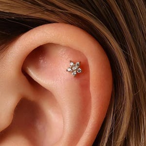May include: Close-up of a gold-colored star-shaped earring with multiple clear gemstones. The earring is set against the backdrop of a person's ear, with visible skin and hair.