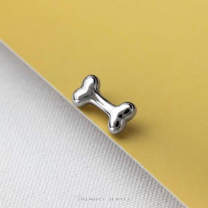 May include: A small, polished silver-toned stud earring shaped like a dog bone. The piece features a smooth, reflective metallic surface with rounded edges and a central indentation. The text SHEMISEI JEWELS is printed at the bottom of the image.