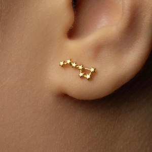 May include: A gold-colored ear cuff featuring a constellation design. The cuff is shaped like a series of connected stars, creating a celestial theme. The jewelry is worn on the ear, showcasing its delicate and detailed craftsmanship.