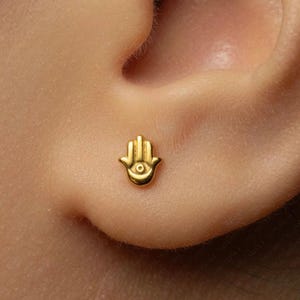 May include: A small gold-toned stud earring shaped like a Hamsa hand. The earring features a smooth, polished surface with a central circular cutout detail, designed to be worn as a single piece of jewelry on the earlobe for a subtle appearance.
