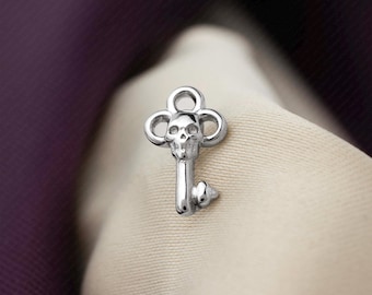 May include: A silver-toned metal charm shaped like a key with a skull integrated into the bow. The top of the key features a circular loop for attachment, while the bottom end has a small, pointed bit. The text SHEMISLI JEWELS is visible.