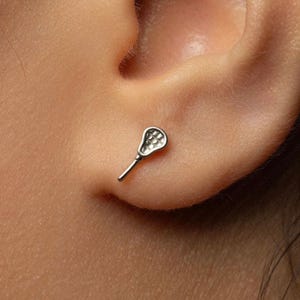 May include: A small silver-toned stud earring shaped like a lacrosse stick. The earring features a long, thin handle and a rounded head with a grid-like mesh pattern. The piece is positioned against the skin of an earlobe, showing its metallic finish.